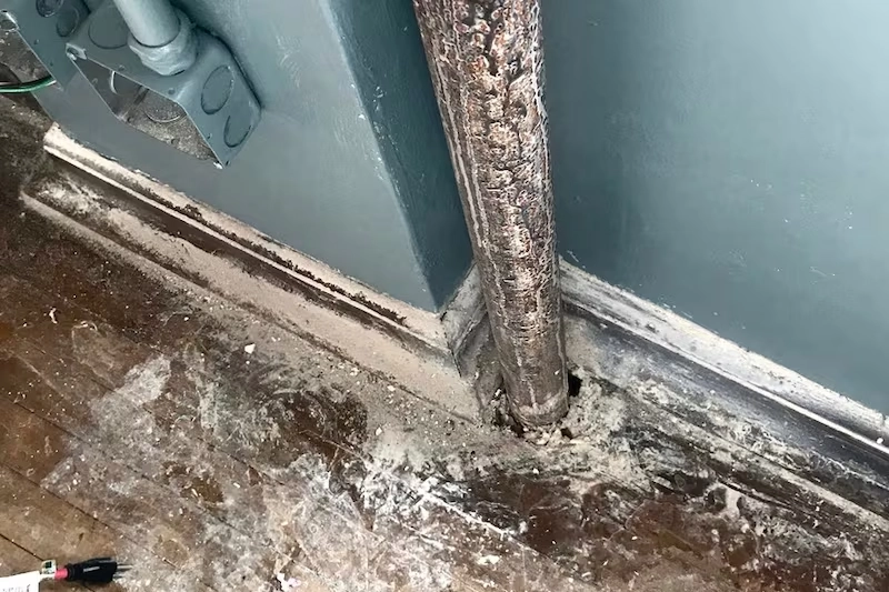 Corner of a deteriorating classroom with a metal pipe emerging from the floor, surrounded by cracked, peeling paint and debris on a wooden floor and baseboard, showing signs of damage and decay.